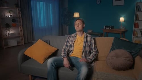 A Young Man Sits on the Sofa in the Evening in the Room and Watches TV The Man Doesn't Like the Show