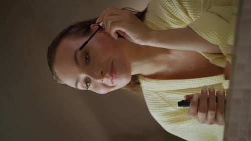 Vertical Screen A Woman in a Yellow Robe Applies Mascara to Her Eyelashes While Looking in a Mirror