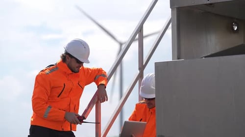 Two engineers in orange jackets and hard hats inspect a wind turbine on a platform