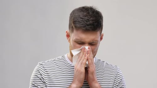 Young man blowing nose with tissue suffering from cold allergy or rhinitis