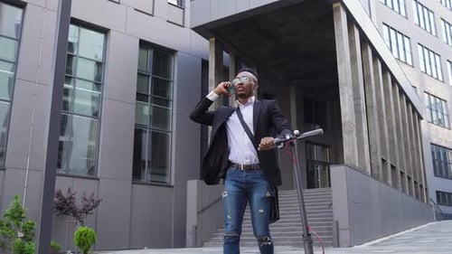 Man in sunglasses walking with own e-scooter near office building and talking on phone