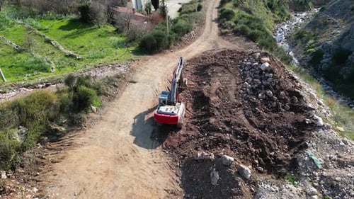 A Drone Flies Over an Excavator Standing Near a Mountain River Next to a Pile of Soil and Stones on