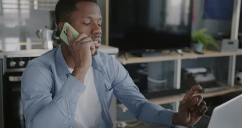 Trabajador independiente afroamericano que habla por teléfono móvil y usa una computadora portátil trabajando en la cocina en