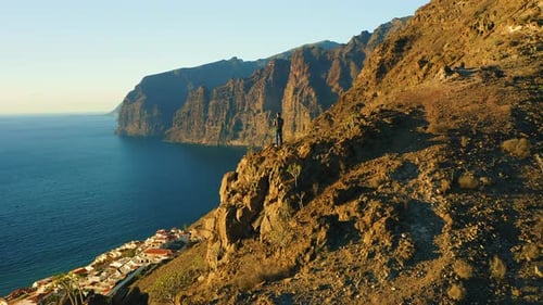 Man Backpacker Standing on Edge of Mountain Cliff with Rocky Beach and Ocean at Sunset