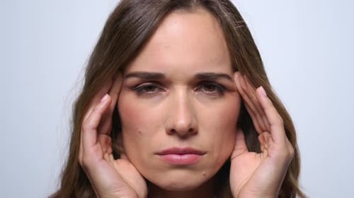Exhausted brunette woman with migraine touches head on blue studio background