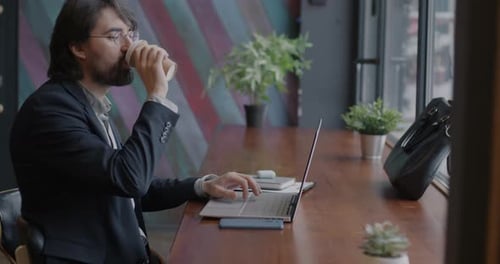 Middle Eastern Businessperson Working with Laptop Computer and Drinking Coffee at Table in Cafe
