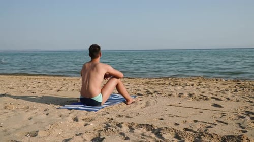 Gebräunter schlanker kaukasischer Mann sitzt auf einem Strandtuch am leeren Sandstrand in der Nähe des Meeres