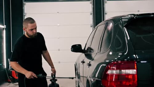 A male car wash employee applies car wash detergent to a black luxury car using spray gun