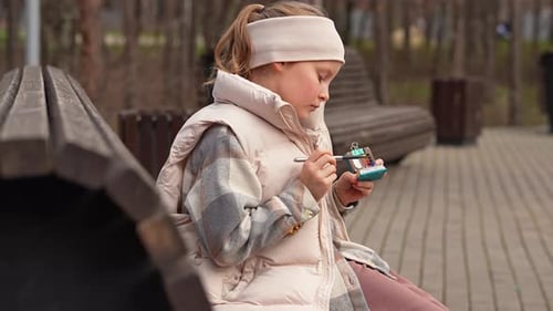 Caucasian School Girl is Sitting on the Bench in Public Park and Drawing in Little Sketchbook