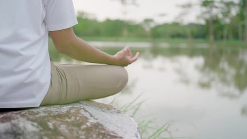 Man meditates on a large rock by the water. He sat cross-legged amidst beautiful nature