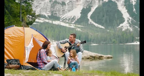 A happy family camping at the lake, playing the guitar and singing a song together in front of the
