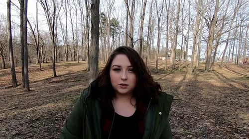 A pretty teenage female wearing a green coat strolls through the park with a serious look on her fac
