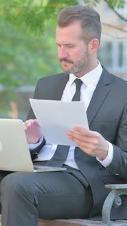 Focused Businessman Analyzing Documents with Laptop Outdoors