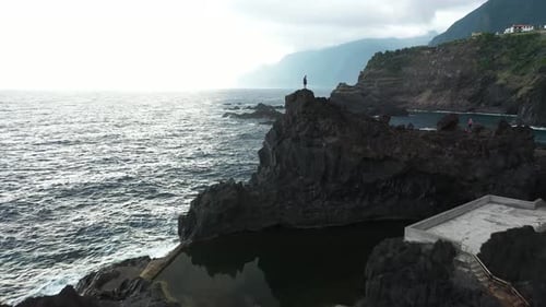 Drone clip flying by a young and fit man, standing on the top of a cliff next to Sexial Natural Pool