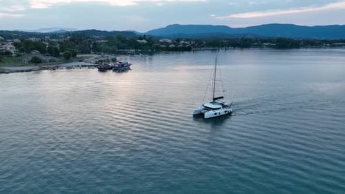 Aerial view of catamaran yacht sailing calmly towards harbor entrance at sunset with mountains