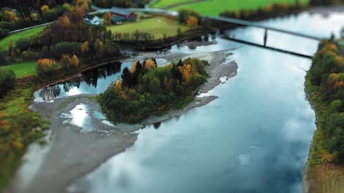 A bridge crosses over a winding river, surrounded by fields and trees dressed in early autumn colors