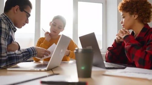 Team of Office Workers Sit at Table with Documents and Laptops and Discuss Work Plan