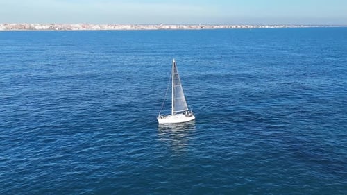 Solitary Sailboat Cruising on Tranquil Blue Sea Against Coastal City Backdrop.