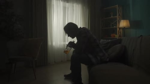 A Devastated Young Man Sits in a Room in the Dark with a Bottle of Alcohol and Drinks From His