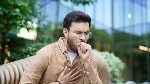Handsome businessman coughing sitting on bench on street near business office building. Sick male