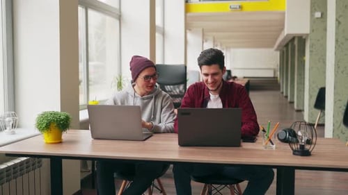 Zwei Hipster-Mitarbeiter tauschen Ideen zum Projekt aus und sitzen mit Laptops im geräumigen Großraumbüro