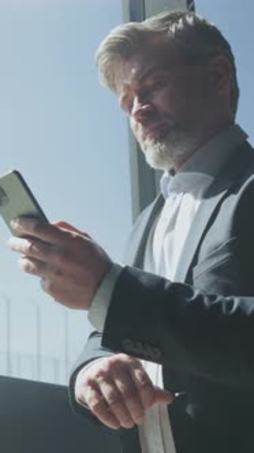 Man in suit uses smartphone, standing by window