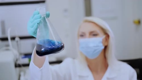 Chemiker mit Gesichtsmaske beobachtet blaue chemische Lösung in einer Laborflasche