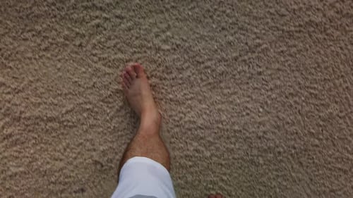 Bare feet of man in swimming trunks walking on a beach. Shade covers sandy shore with legs of person