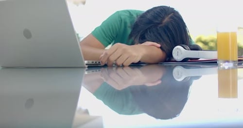 A young Asian male college student is resting head on table beside laptop