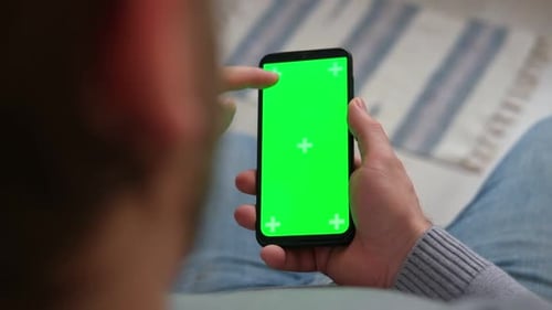 Young Man Sitting at Home Holding Smartphone Green Mockup Screen in Hand Male Person Using Chromakey