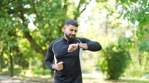 Bearded male runner jogging in an urban city park and looking smart watch. Handsome athlete using