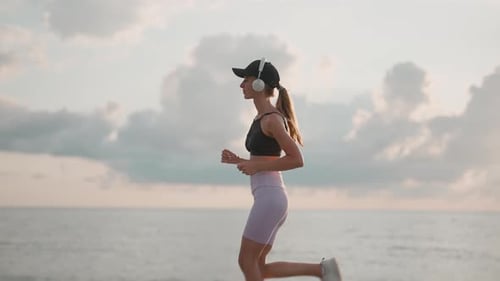 A Young Woman Runner is Listening to Music in Earphones and Training on a Sea Beach