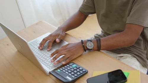 African man placing hands on laptop typing budgeting calculations at wooden table with calculator