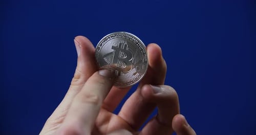 Close-up of hands holding and inspecting a silver Bitcoin against a solid blue background