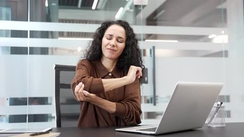 Businesswoman suffering from elbow pain while sitting at a desk at a workplace in a business office.