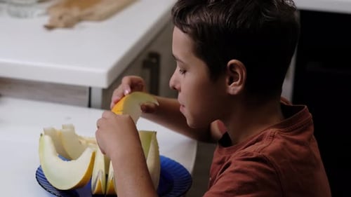Assis à une table de cuisine, un garçon vêtu d'une chemise orange déguste une tranche de melon. Cette scène met en valeur le