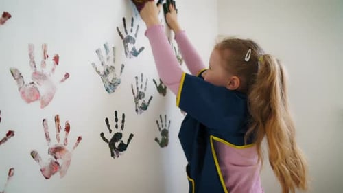 Back view of a fun mischievous baby girl leaving colored hand prints on white wall wall in room.