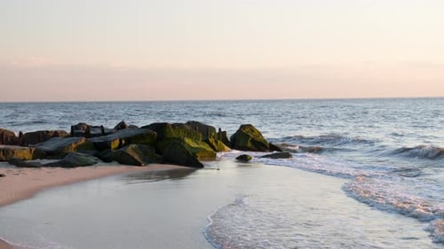 Waves Splash Against Emerald Green Mossy Rocks and Wash Ashore on Clear Evening in Slow Motion