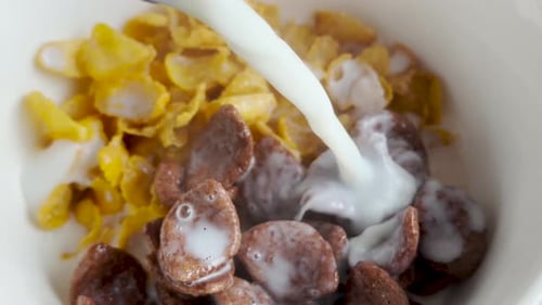 Delicious Breakfast: Pouring Hot Milk Over Corn Flakes and Chocolate Flakes in White Bowl - Close Up