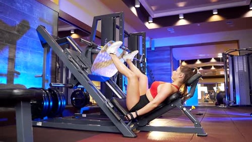 Strong young woman in sportswear using weight machine for legs at modern gym. Attractive woman