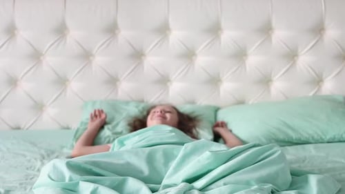A little girl sits on a bed in pajamas and smiles at the camera, and than falls on the bed