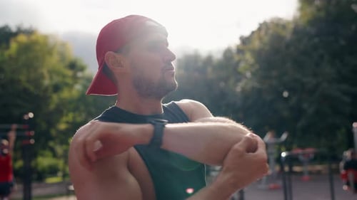 Young Man Stretching Arms In Park Wearing Red Baseball CapPreparing For Workout
