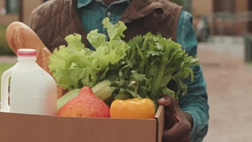 Man holding a box filled with fresh milk, bread, and green produce