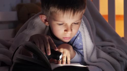 Closeup of boy holding flashlight reading book in bed