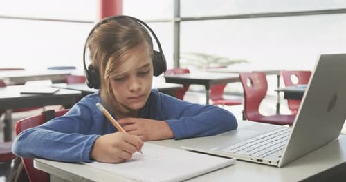 In school, boy wearing headphones writing in notebook, using laptop on desk