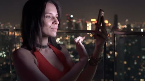 Young, Elegant Woman Texting On Smartphone Standing On Terrace At Night