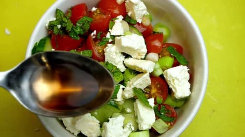 Top down slow motion of a hand pouring cider vinegar over a greek style salad with a yellow backgrou