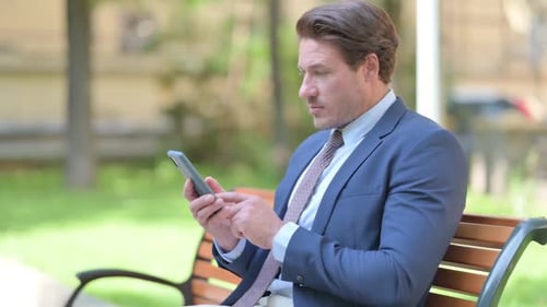 Side View of Businessman using Smartphone sitting Outside Office