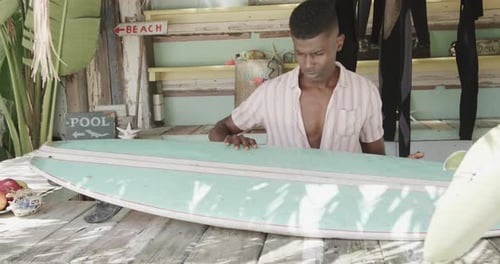 African american man preparing surfboard on the counter of surf rental beach shack, slow motion