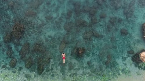 Young Caucasian man swims out from the shore into open turquoise water, high overhead aerial